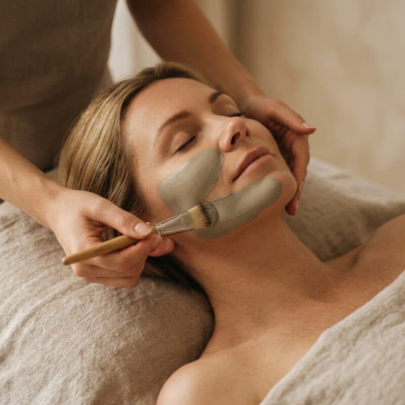 A grey clay mask being brushed along the jaw of a woman resting with her eyes closed.