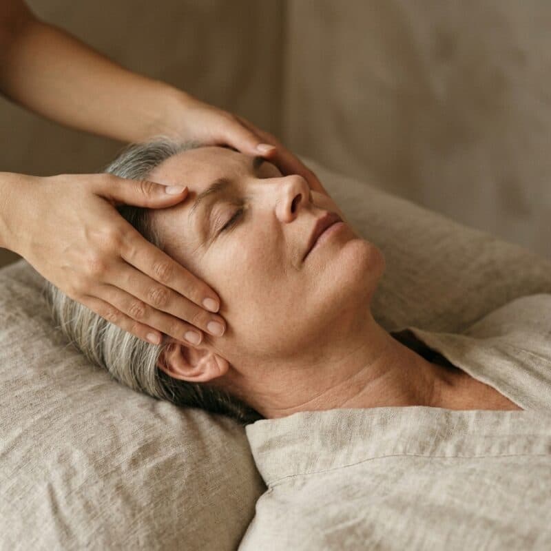 Two hands resting either side of the forehead of a silver-haired woman on a linen pillow.