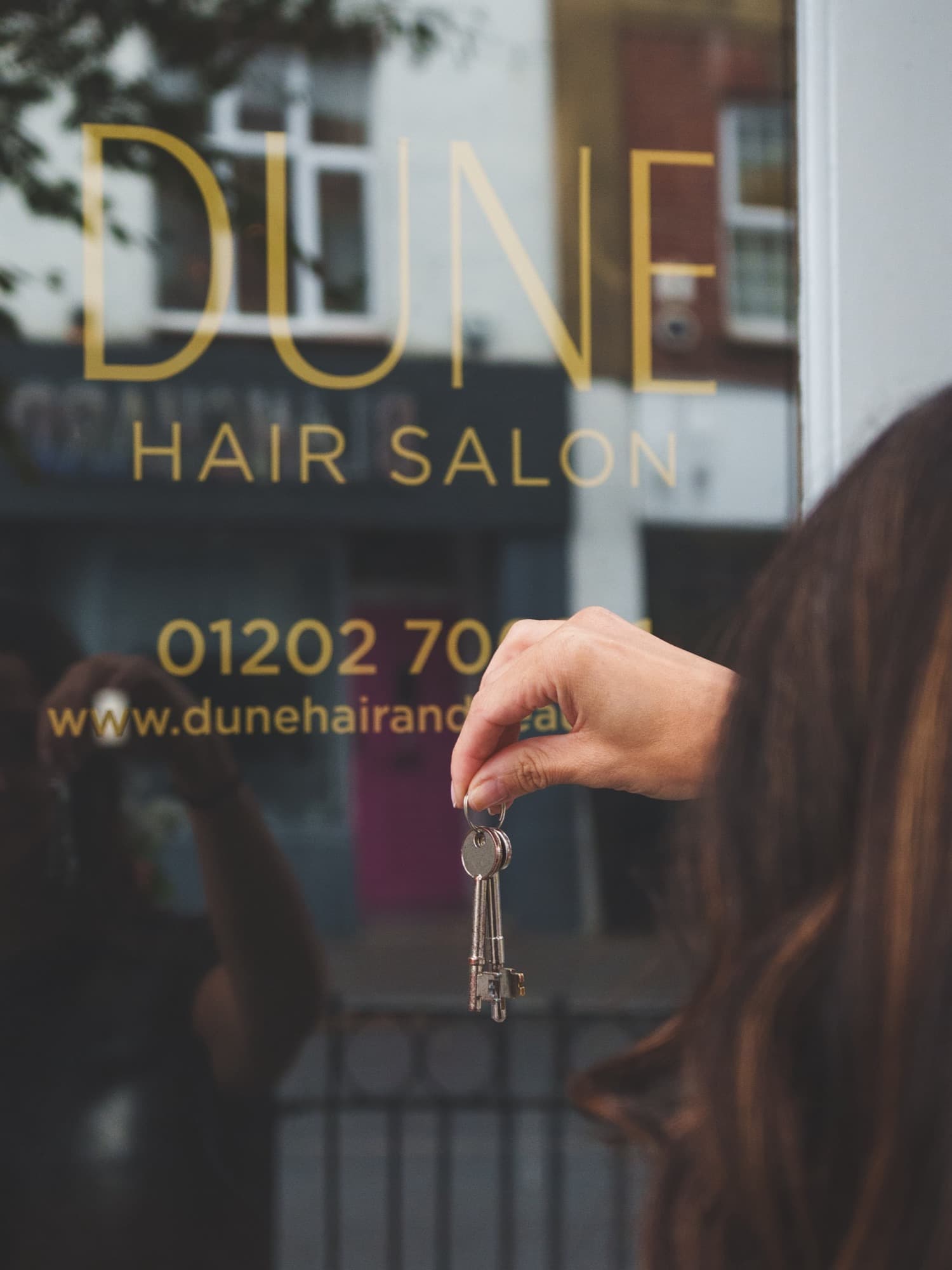 A hand holding a set of keys up against a shop window lettered in gold with Dune Hair Salon, the parade opposite reflected in the glass.