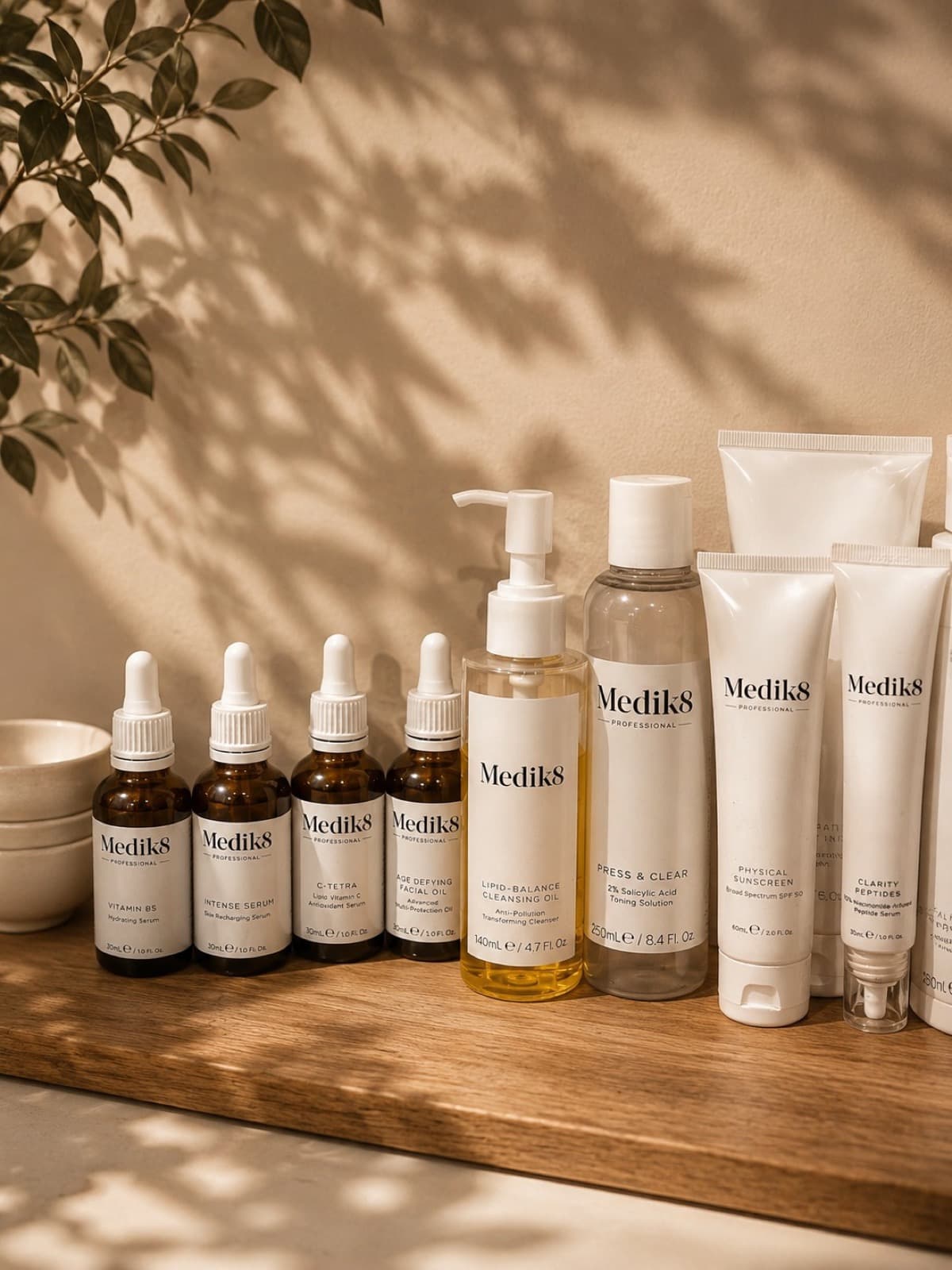 A row of Medik8 serums and cleansers on the wooden counter, leaf shadows across the wall behind.