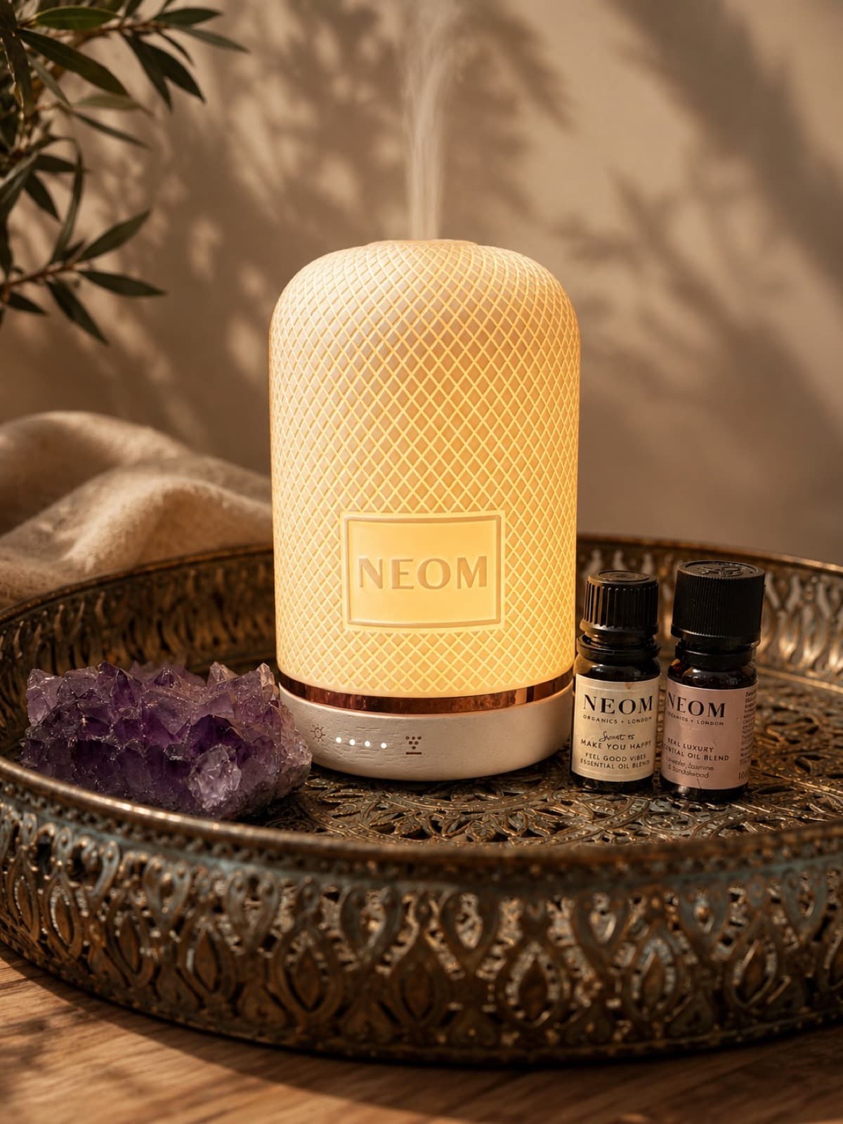 A lit NEOM diffuser breathing a thread of steam, two essential oil bottles and a piece of amethyst on a brass tray.