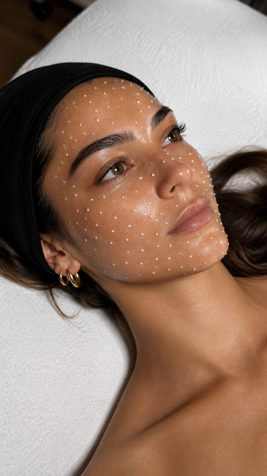 A woman lying back with a dark headband, fine white dots of serum dotted across her cheek and brow, gold hoops at her ear.