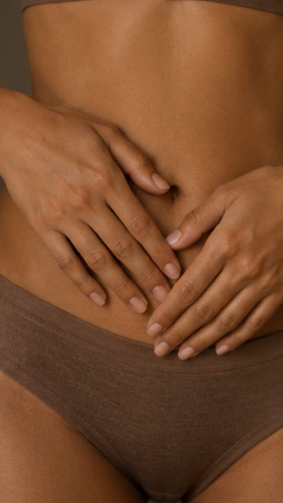 Two hands resting gently across a bare stomach above soft brown underwear.